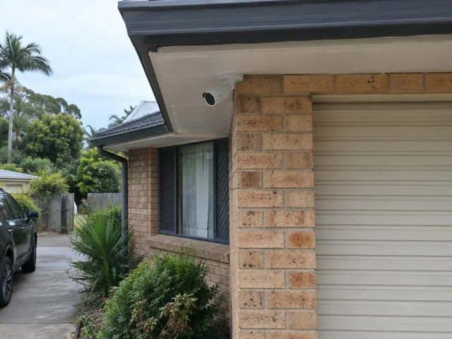 White CCTV camera mounted under a brick house eave beside a garage in Highland Park